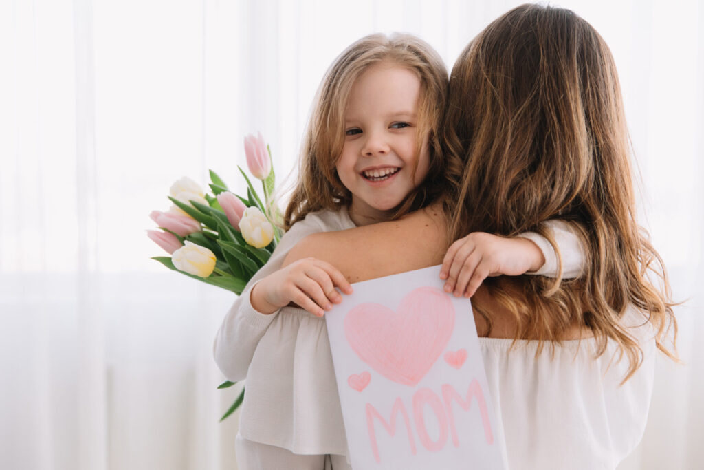 Smiling little girl hugging her mother while holding a handmade Mother&rsquo;s Day card and tulips indoors.